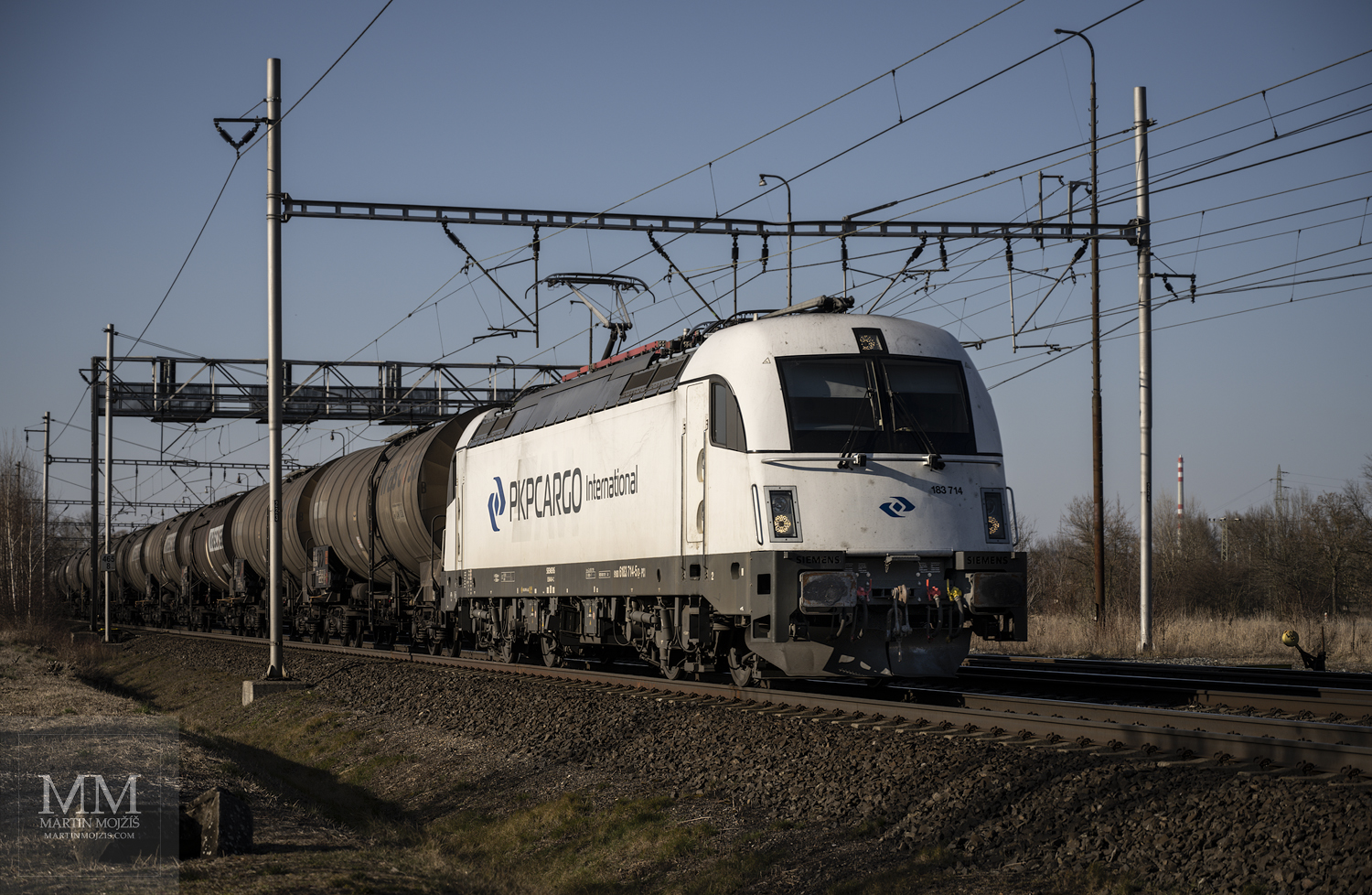 Bílá elektrická lokomotiva Siemens Taurus 183 714 PKP Cargo International v čele nákladního cisternového vlaku.