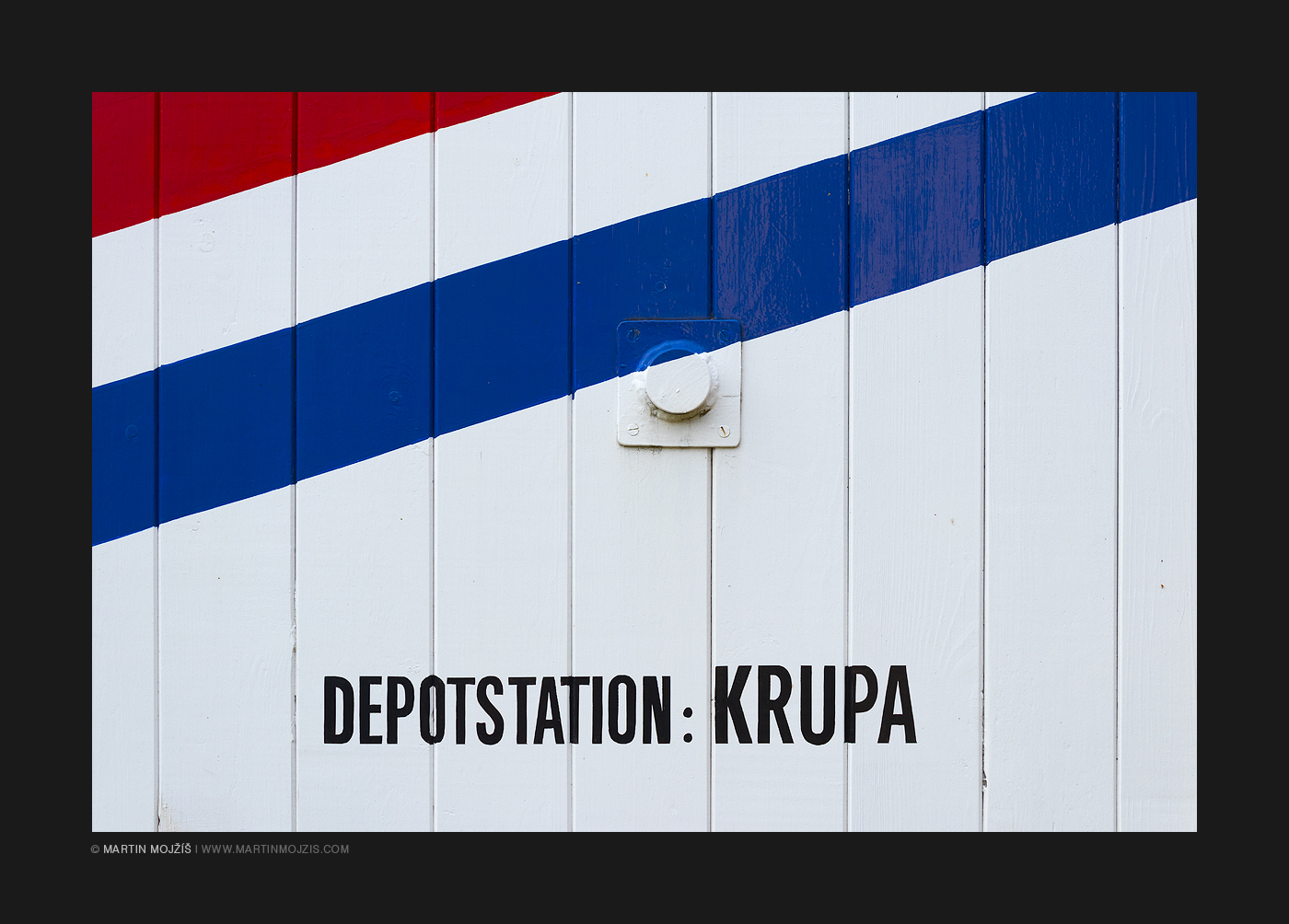 Bíle natřená dřevěná stěna vagonu, červený a modrý pruh. Nápis Depotstation: Krupa. Železniční muzeum v Lužné u Rakovníka.