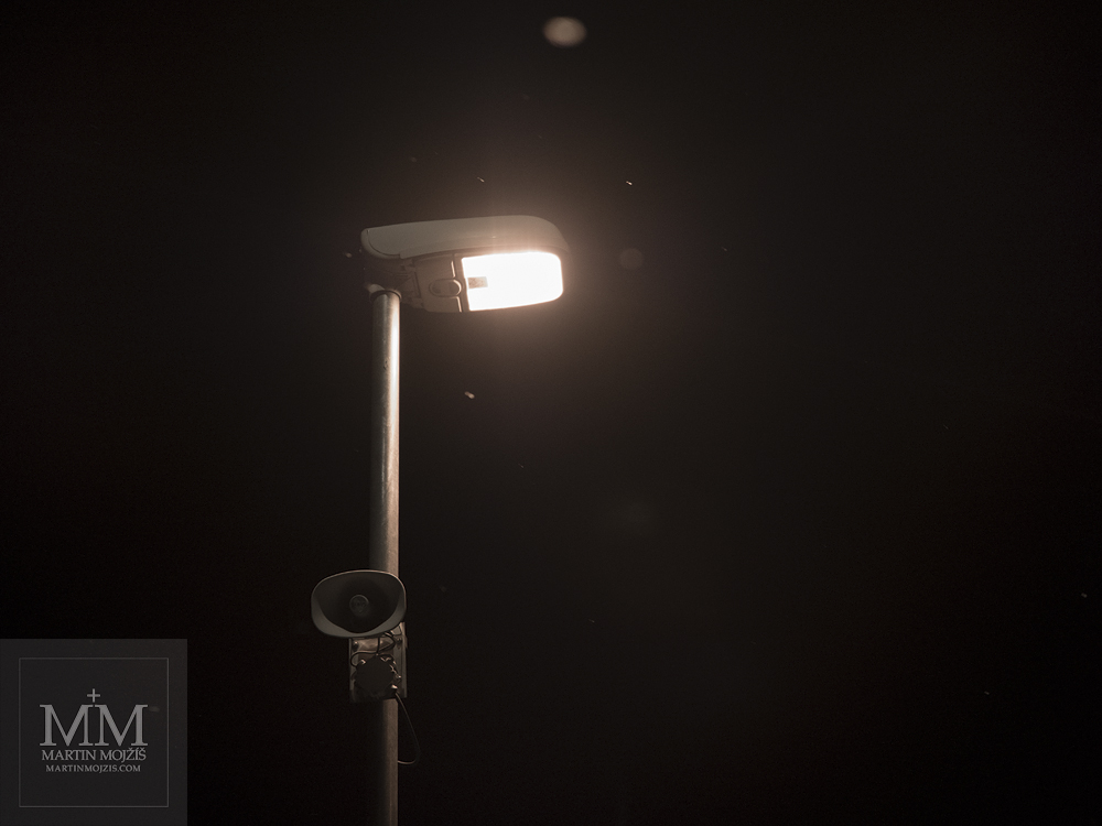 Svítící lampa na železničním nástupišti, padající sníh. Fotografie zhotovená objektivem Olympus M. Zuiko digital ED 25 mm 1:1.2 Pro.