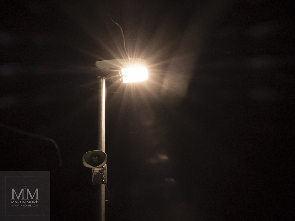 Svítící lampa na železničním nástupišti, padající sníh, velmi silné odlesky. Fotografie zhotovená objektivem Olympus M. Zuiko digital ED 25 mm 1:1.2 Pro.