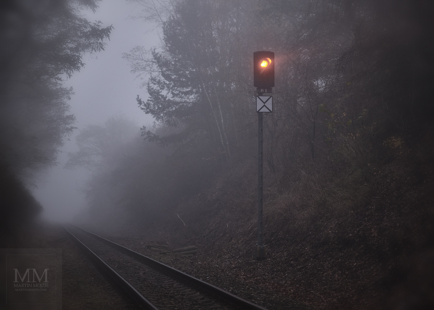 Železniční světelné návěstidlo (semafor) u jednokolejné trati svítící v mlhavém soumraku.