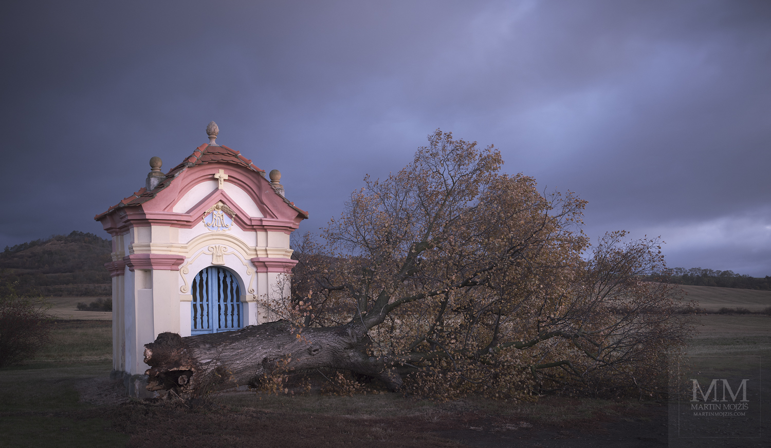 A fallen tree in front of a chapel in a landscape. Fine art photograph AFTER A BIG WIND, photographed by Martin Mojzis.