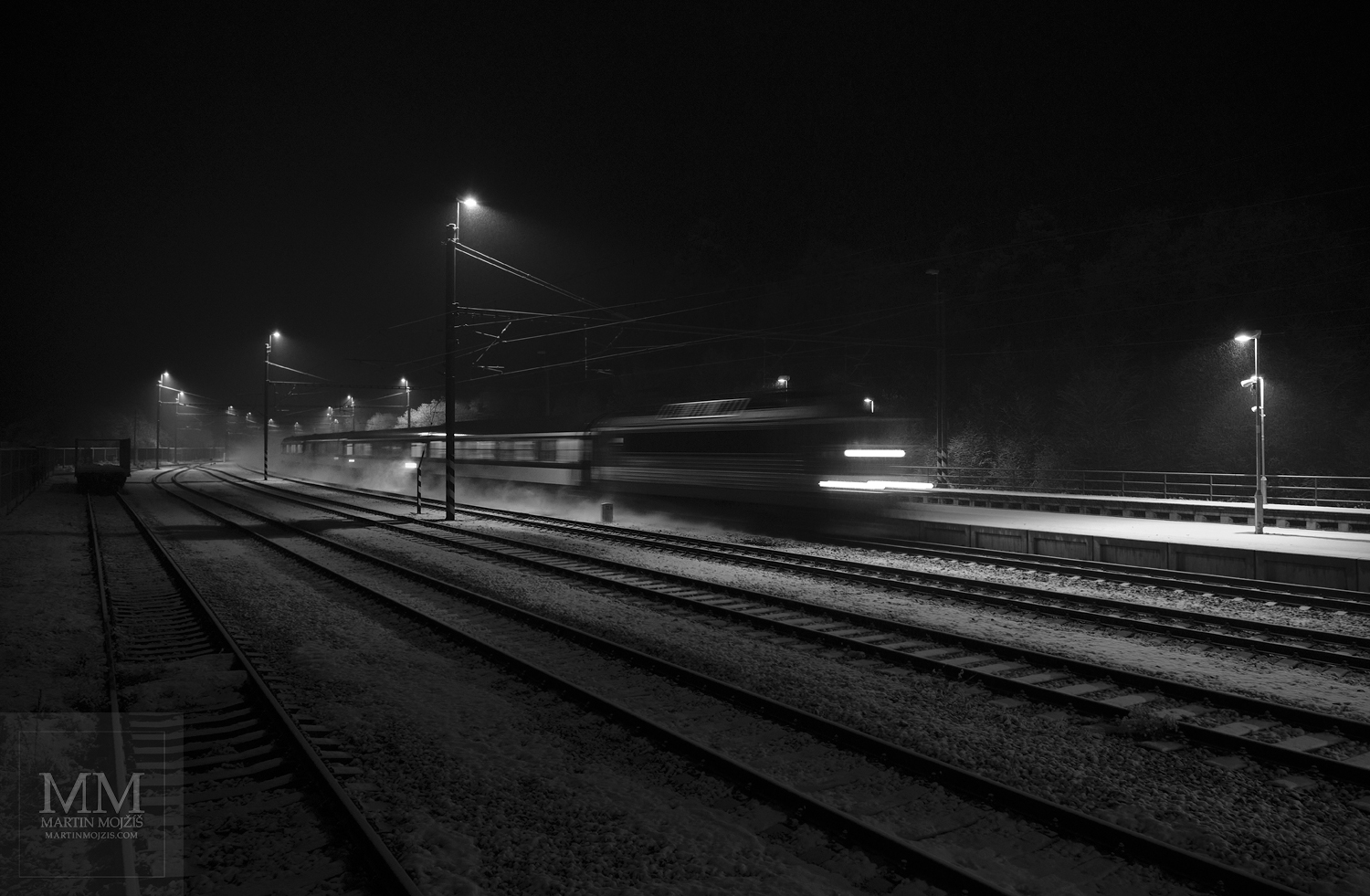 Noční vlak jedoucí po osvětlené zasněžené trati. Umělecká černobílá fotografie s názvem ZASNĚŽENOU NOCÍ I. Fotograf Martin Mojžíš.