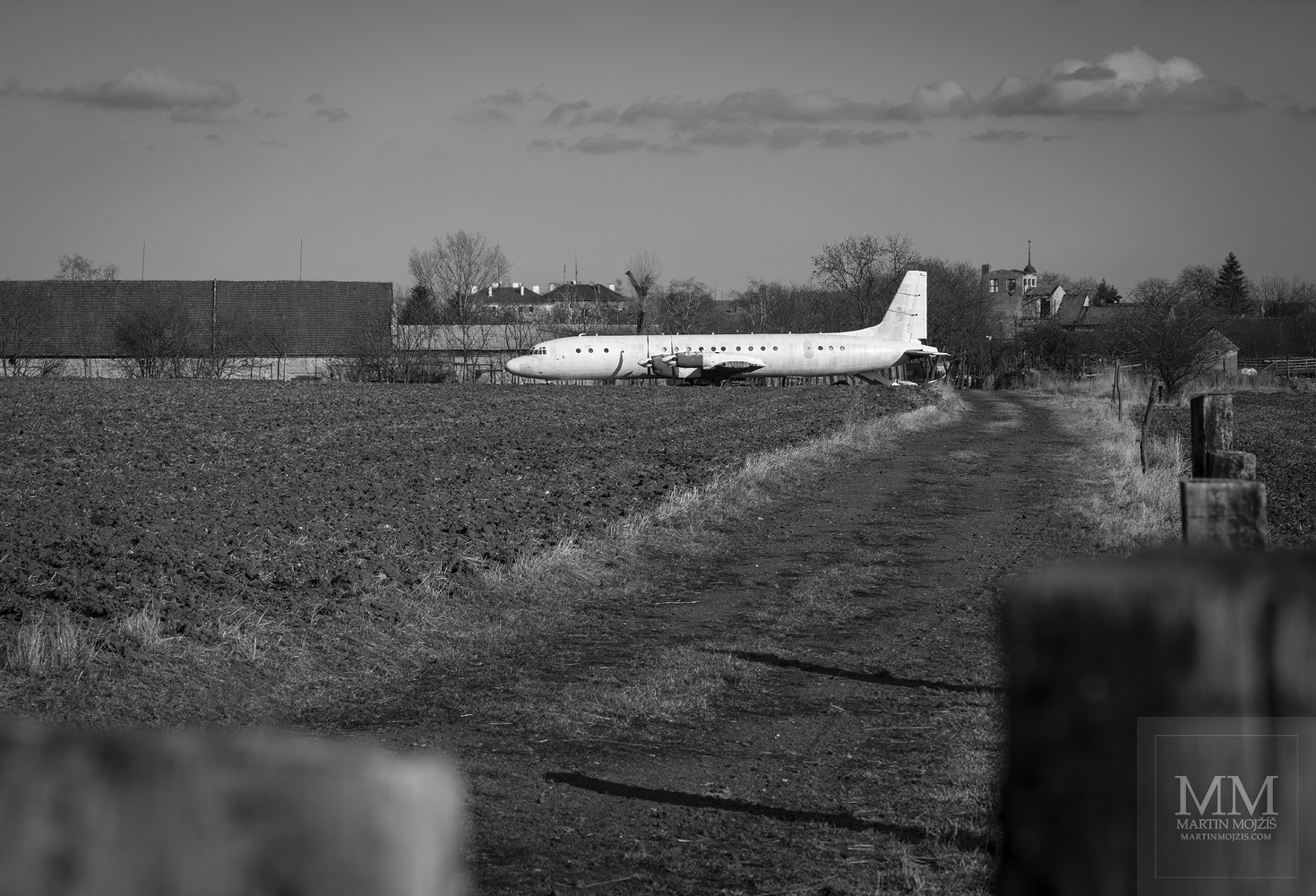 A large white airliner in a field near a farm. Black and white fine art photograph IN THE HALF OF FEBRUARY, photographed by Martin Mojzis.