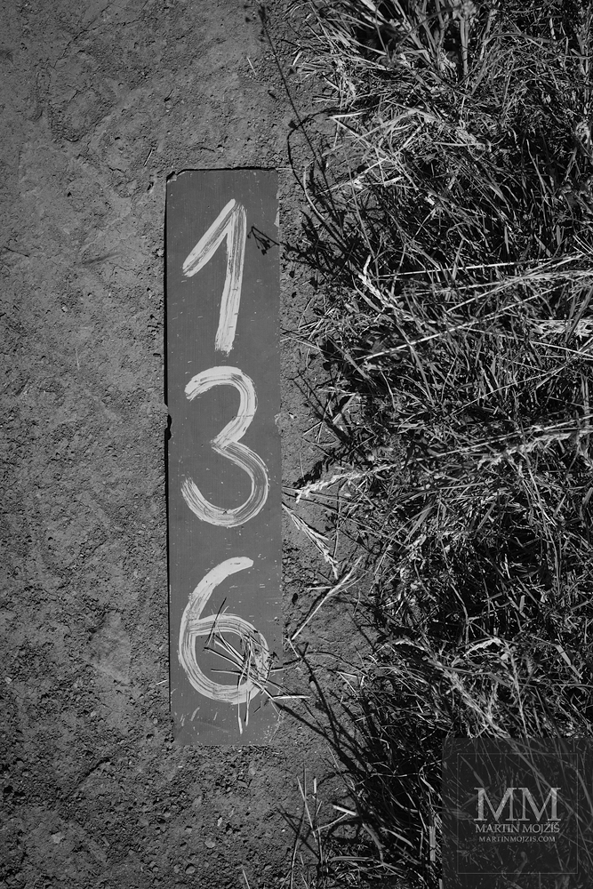 Plechová tabulka s čislem 136 ležící na prašné cestě u pole. Umělecká černobílá velkoformátová fotografie s názvem 136. Fotograf Martin Mojžíš.