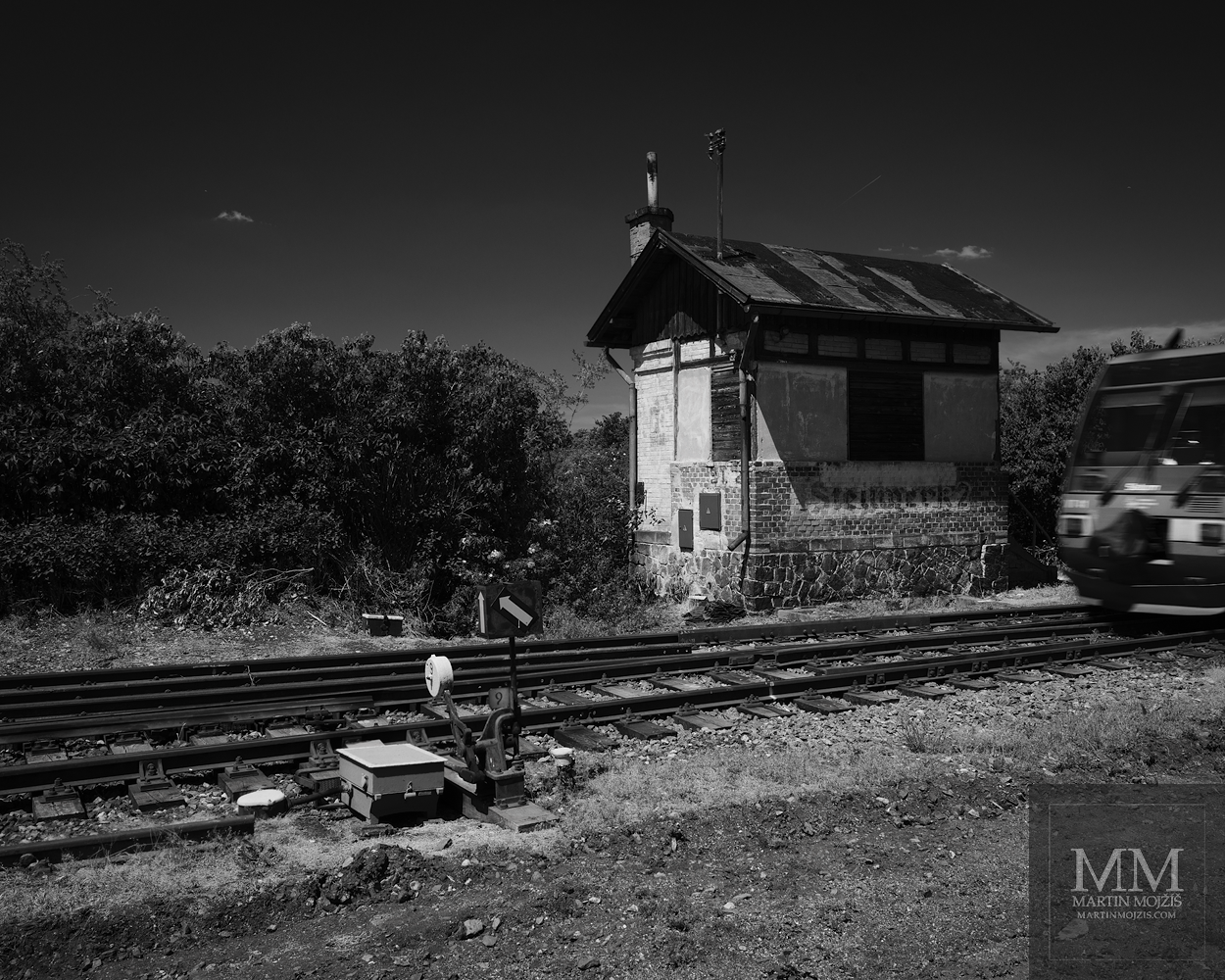 Historický železniční strážní domek, výhybka a projíždějící vlak. Umělecká černobílá velkoformátová fotografie s názvem DEVÁTÁ VÝHYBKA. Fotograf Martin Mojžíš.