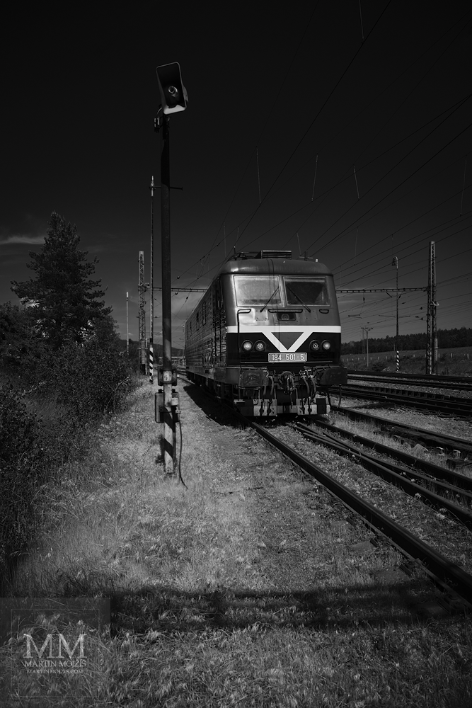 Lokomotiva 184 501-5 na nádraží. Umělecká černobílá velkoformátová fotografie s názvem V ODPOČINKU POD HORAMI. Fotograf Martin Mojžíš.