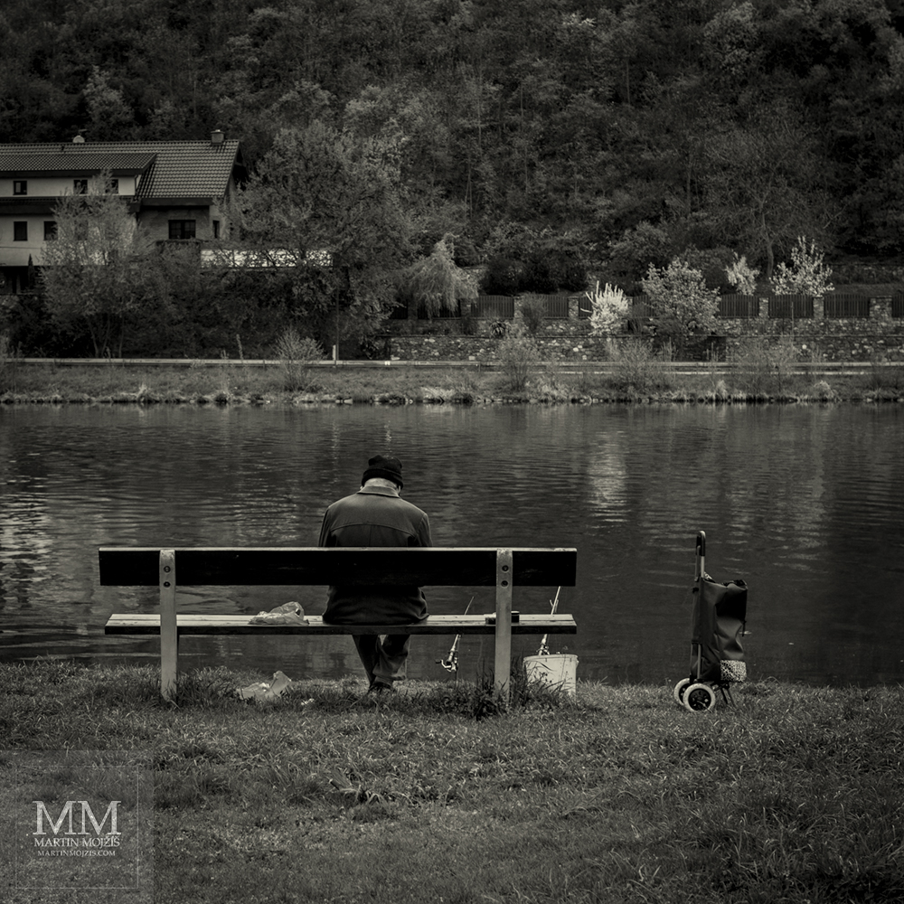 Rybář na lavičce u řeky. Fotografie s názvem ČEKEJ.