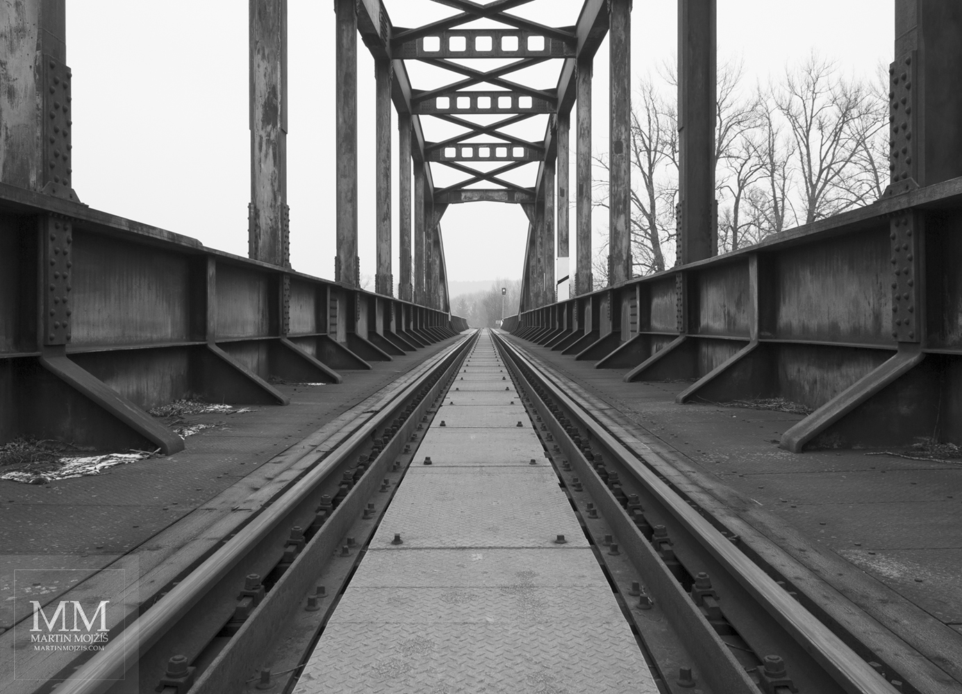 Jednokolejná trať vedoucí po ocelovém mostě. Umělecká černobílá fotografie Martina Mojžíše s názvem AŽ SE BUDE ŠEŘIT.