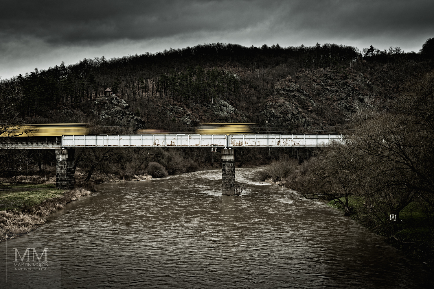 Pohybově rozostřený žlutý vlak jede po mostě přes řeku, tmavé mraky.