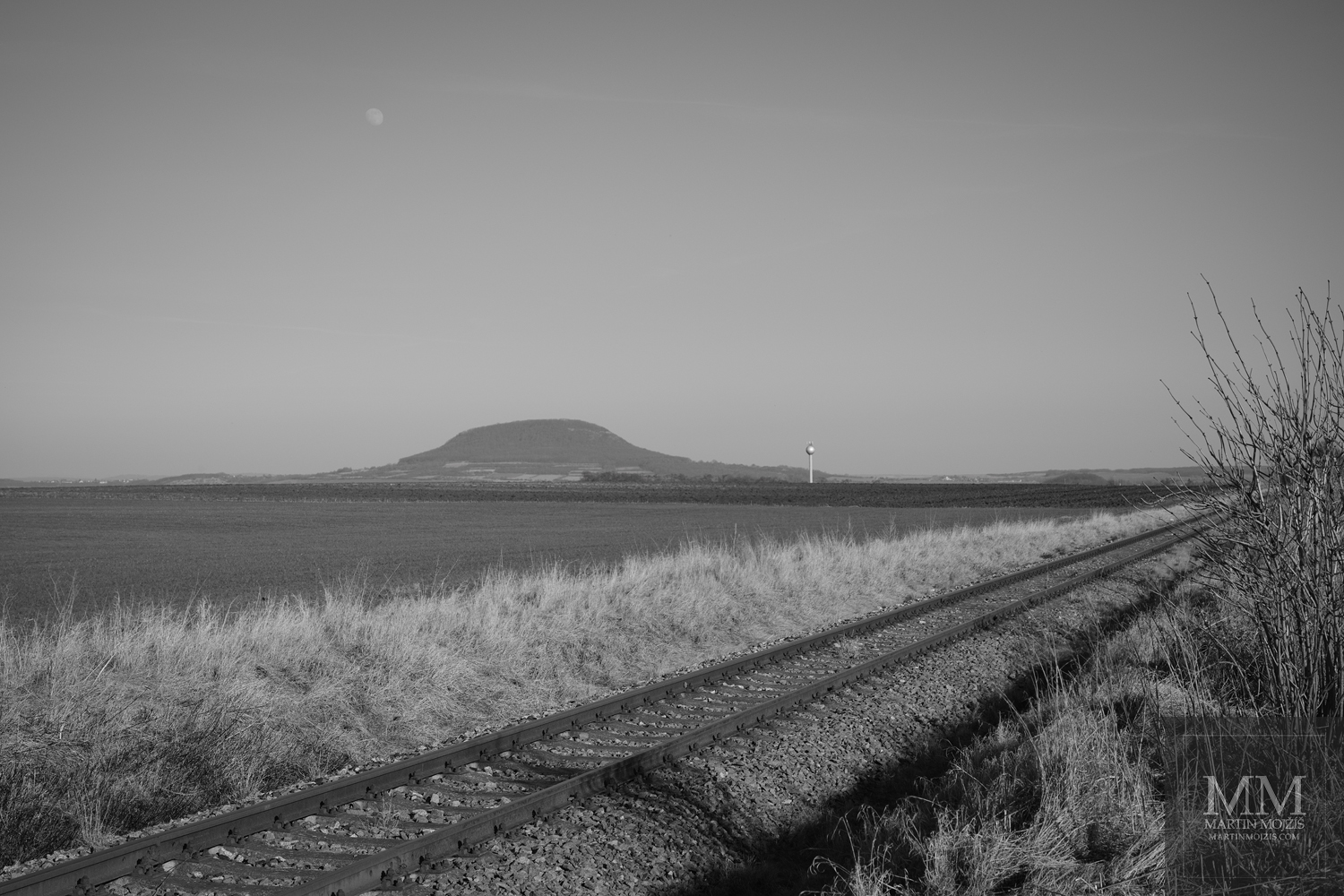 Železniční trať nedaleko hory Říp. Velkoformátová černobílá umělecká fotografie krajiny s názvem KDE JEZDÍ VLAKY. Fotograf Martin Mojžíš.